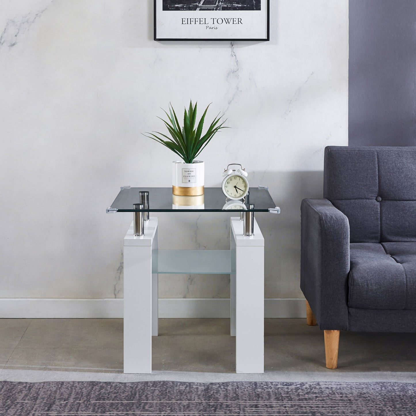 A Clear Glass Top Side Table, Modern Design For Home, stands by a gray sofa with a potted plant and alarm clock. Above the table, a framed Eiffel Tower print decorates the white and gray wall.
