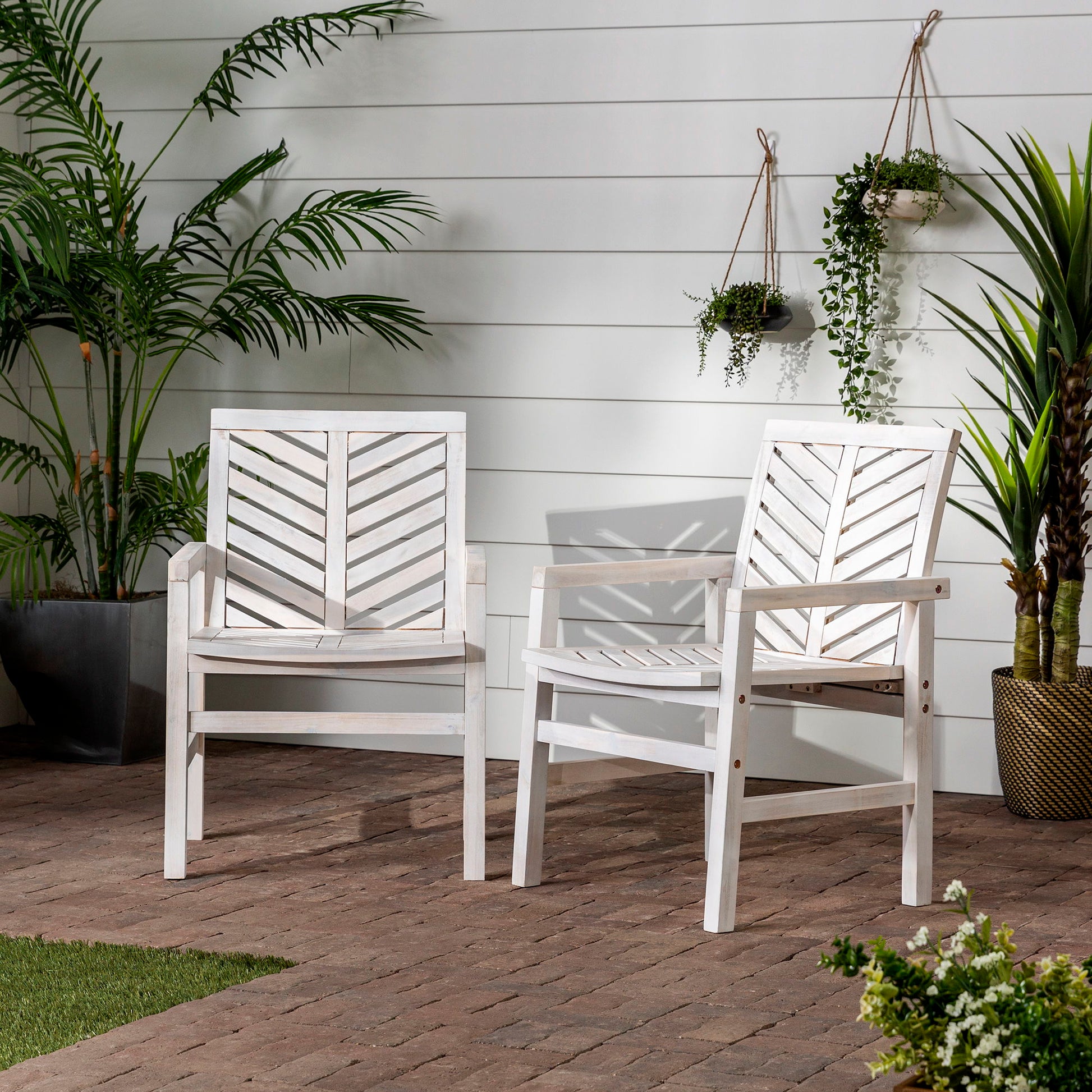 The Modern 2 Piece Chevron Patio Chairs, finished in white wood with chevron-patterned backs, are set on a brick patio surrounded by green potted plants and hanging planters beside a white paneled wall.