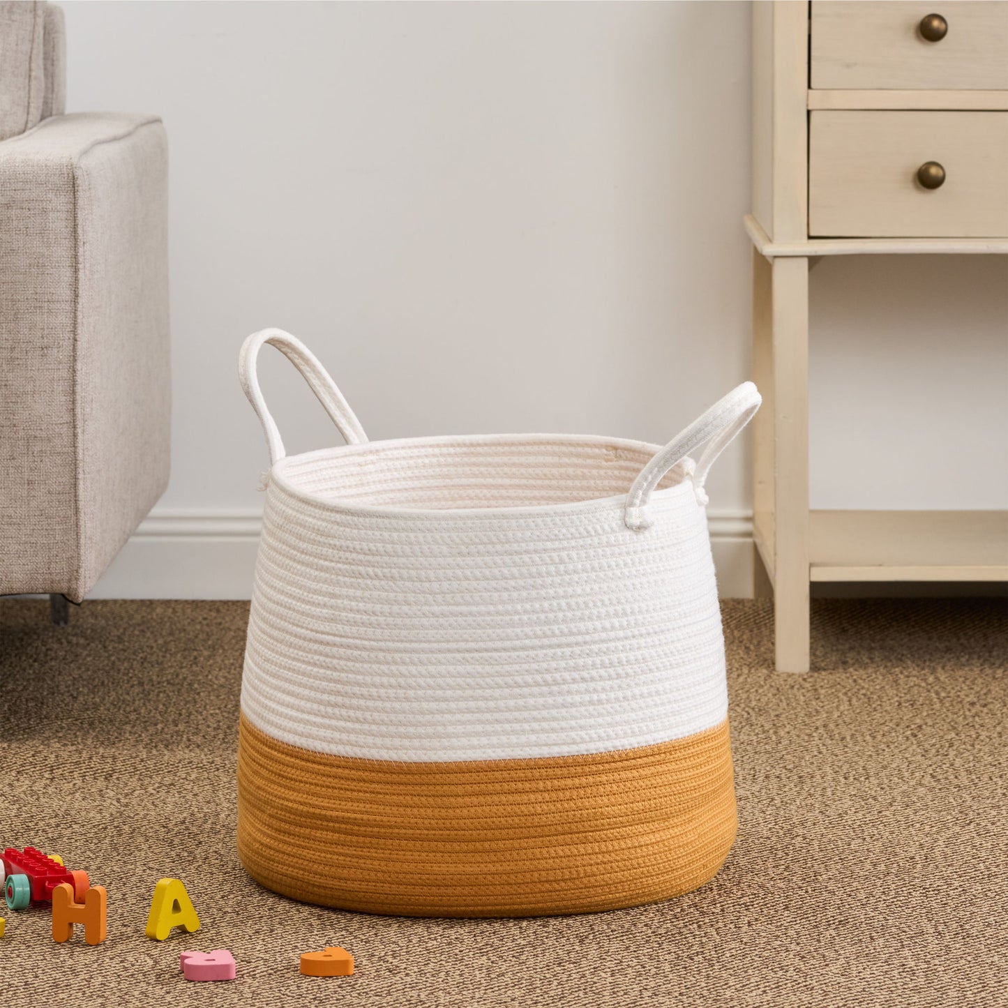 The Francis - Cotton Round Laundry and Storage Basket in white and mustard yellow, featuring handles, is placed on a carpeted floor near a beige sofa and light wood dresser with colorful toy blocks scattered nearby.