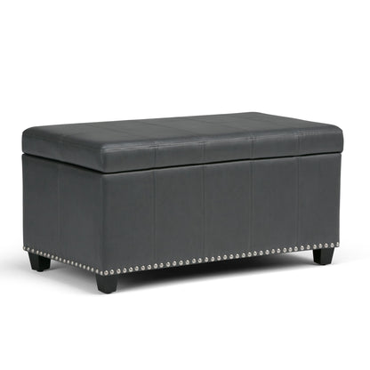 The Amelia Transitional Storage Ottoman Bench features a rectangular black leather design with a cushioned lid, decorative stitching, silver nailhead trim at the base, and short wooden legs.
