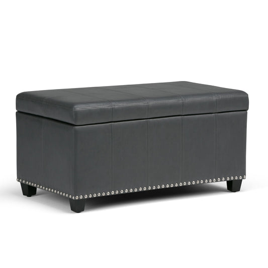 The Amelia Transitional Storage Ottoman Bench features a rectangular black leather design with a cushioned lid, decorative stitching, silver nailhead trim at the base, and short wooden legs.