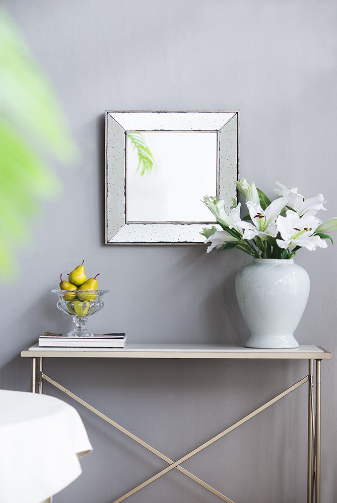 A minimalist console table sits against a light gray wall with the Distressed Accent Mirror, a traditional style framed wall mirror for living rooms, above it. On the table are a white vase of white lilies, yellow pears in a glass bowl, and stacked books.