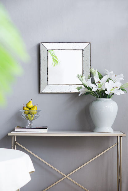 A minimalist console table sits against a light gray wall with the Distressed Accent Mirror, a traditional style framed wall mirror for living rooms, above it. On the table are a white vase of white lilies, yellow pears in a glass bowl, and stacked books.