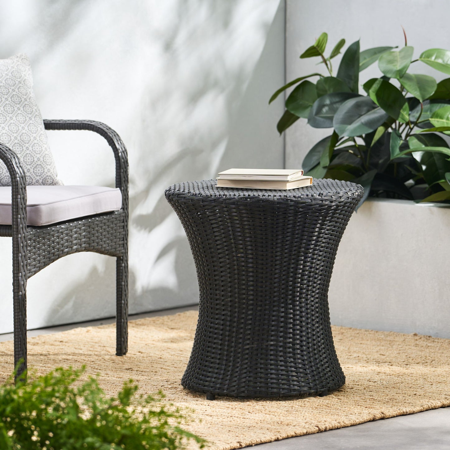 The Thrive Hourglass Rattan Table with no glass top sits on a woven rug beside a matching chair with cushion, while green plants and sunlight enhance the modern outdoor patio’s natural feel.