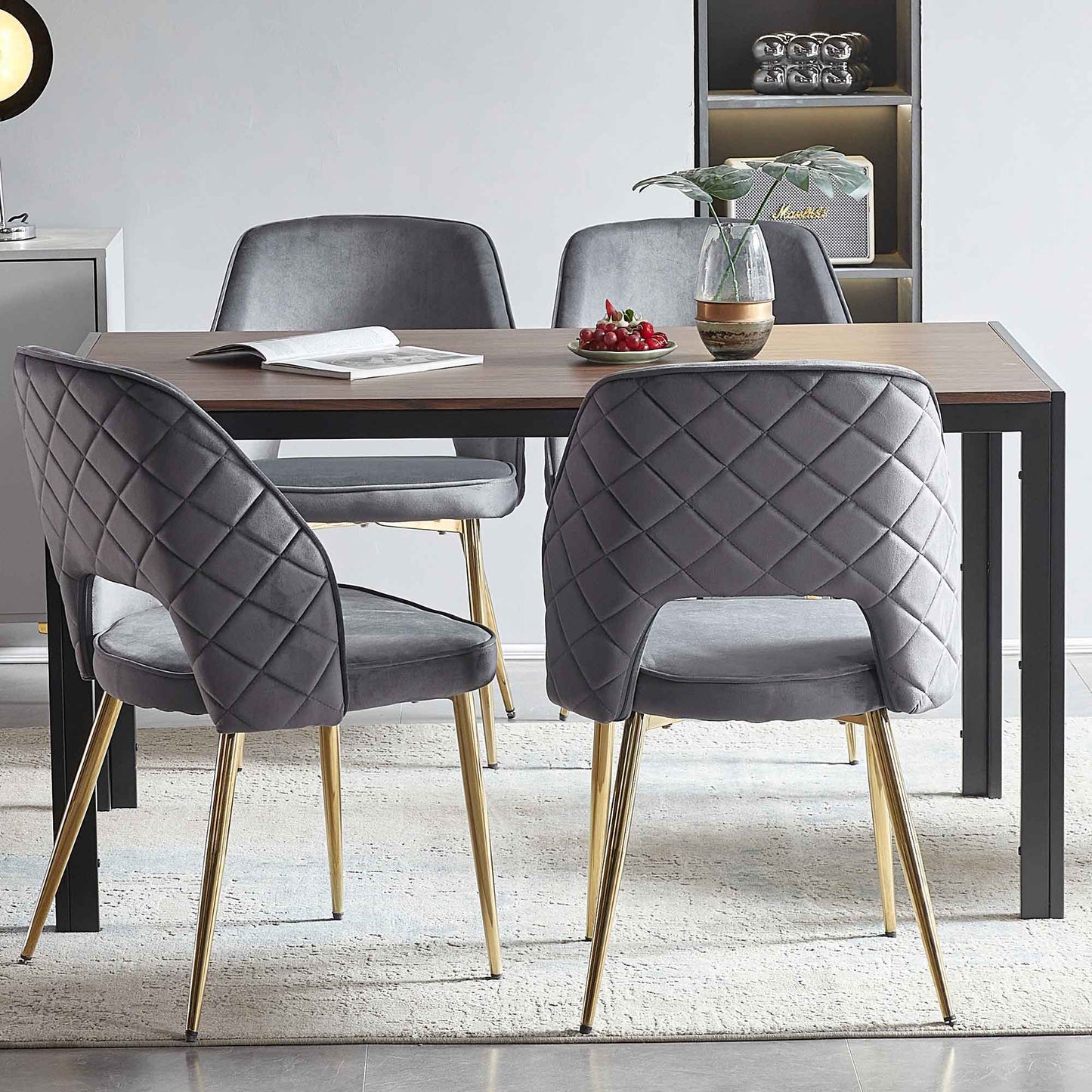 A modern dining area with a rectangular wooden table, four Velvet Dining Chairs With Metal Legs And Hollow Back, a vase with a plant, a plate of cherries, and an open book on the table atop a light rug.