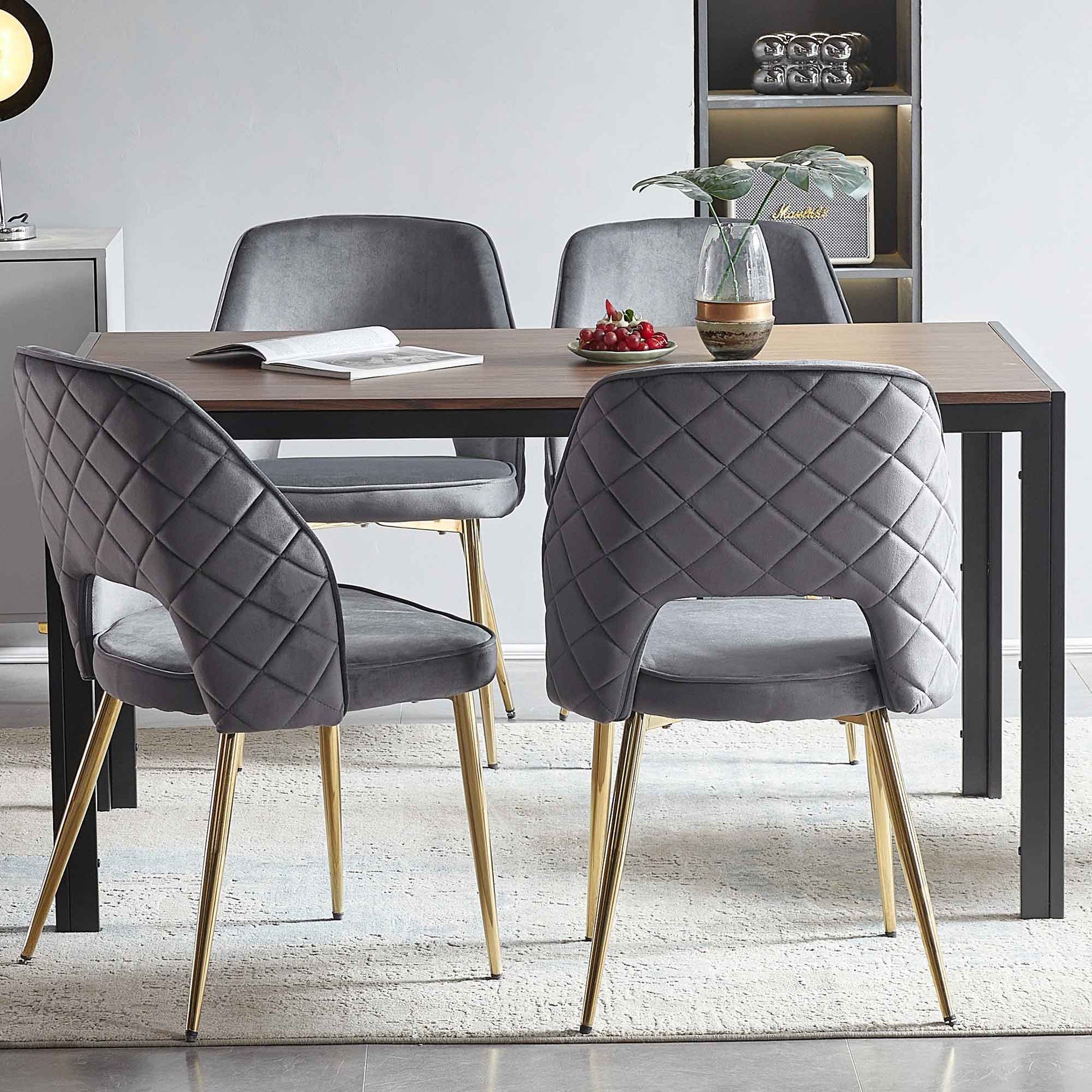 A modern dining area with a rectangular wooden table, four Velvet Dining Chairs With Metal Legs And Hollow Back, a vase with a plant, a plate of cherries, and an open book on the table atop a light rug.