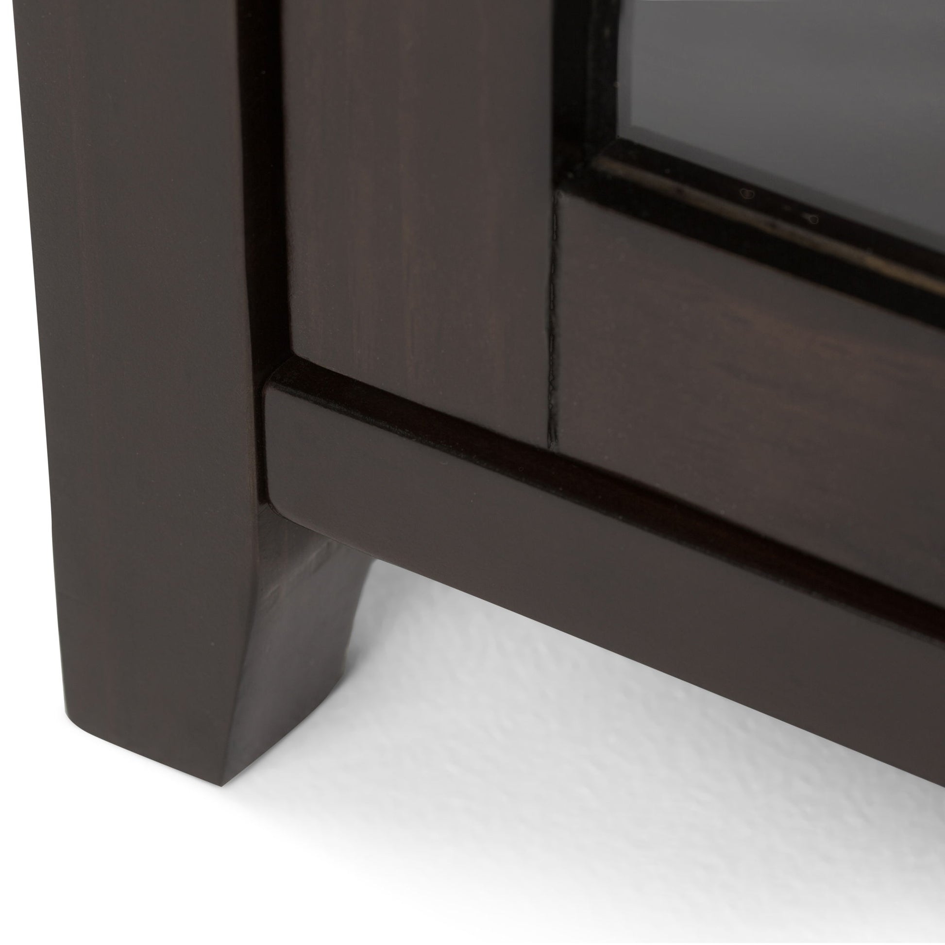 Close-up of the bottom corner of the Cosmopolitan - Handcrafted Medium Storage Cabinet, featuring dark wood and a glass panel, resting on a white surface.