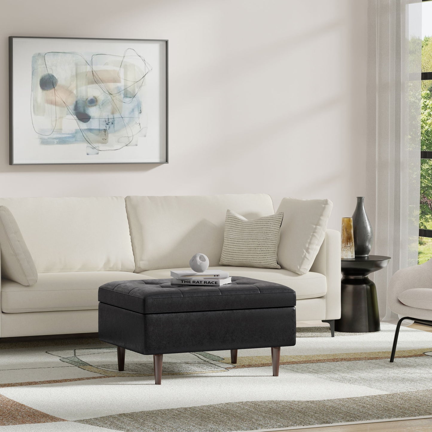 A modern living room with a light-colored sofa, the Shay Large Square Storage Ottoman in black used as a coffee table with books and decor, abstract wall art, a round black side table, and a large window with sheer curtains letting in natural light.