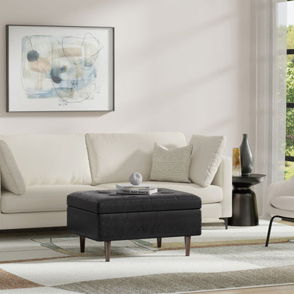 A modern living room with a light-colored sofa, the Shay Large Square Storage Ottoman in black used as a coffee table with books and decor, abstract wall art, a round black side table, and a large window with sheer curtains letting in natural light.