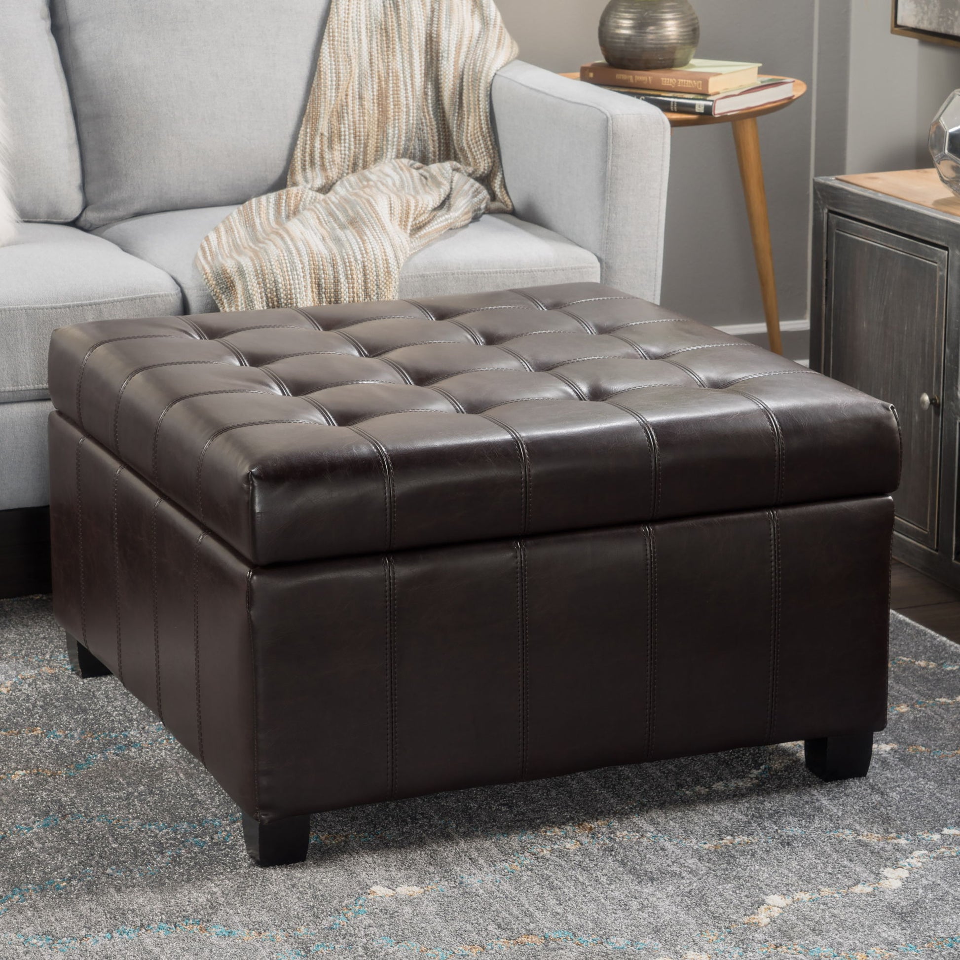 The Isabella - Contemporary Ottoman With Storage in dark brown leather sits on a gray patterned rug near a light gray sofa with a striped blanket, beside a wooden table topped with a lamp and books in the living room.