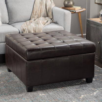 The Isabella - Contemporary Ottoman With Storage in dark brown leather sits on a gray patterned rug near a light gray sofa with a striped blanket, beside a wooden table topped with a lamp and books in the living room.