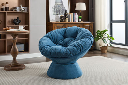 An Oversized Swivel Accent Chair (360° Barrel Papasan) in plush blue sits on a light rug in a modern living room with wooden furniture, a potted plant, and large windows letting in natural light.