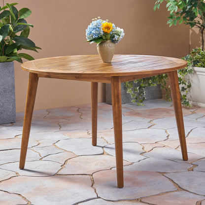 The Stamford - Outdoor Dining Table Modern Design stands on a tiled patio, topped with a vase of blue and yellow flowers and surrounded by green potted plants in the background.