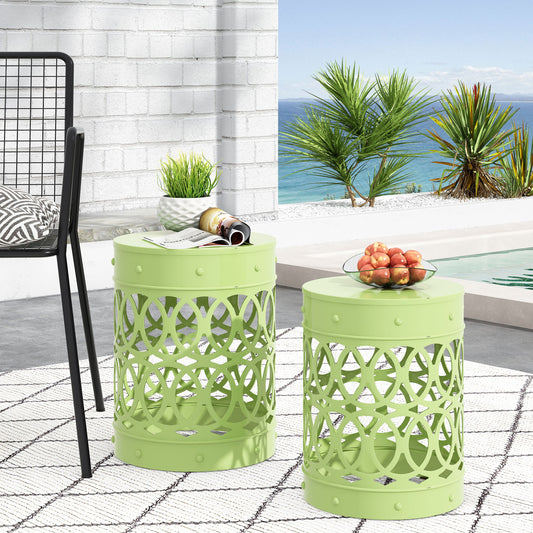 Two Outdoor Metal S/2 End Tables in light green with geometric cut-outs are set on a white patterned rug by a pool. One displays apples, the other magazines and a plant. Palm trees and the ocean appear in the background.