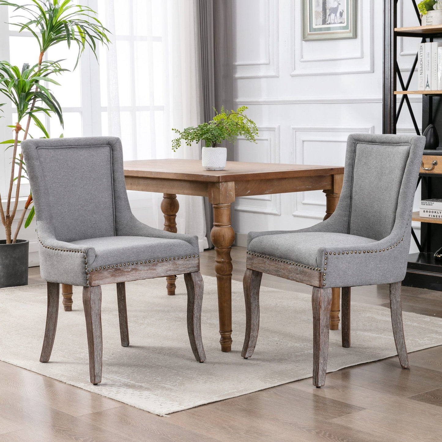 A set of two Ultra Side Dining Chairs with thickened fabric and neutral solid wood legs, featuring bronze nail heads, are positioned by a square wooden table in a bright, modern room with white walls and a large window.