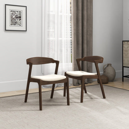 A set of two Dakota Mid-Century Modern Velvet Dining Chairs sits on a light rug in a bright, minimal room with large windows, neutral curtains, wall art, and ceramic vases in the background.