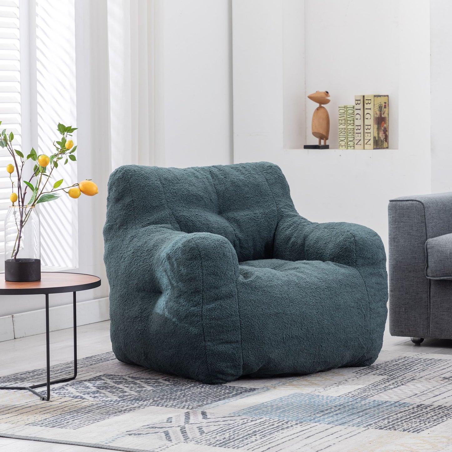 A Soft Teddy Fabric Tufted Foam Bean Bag Chair sits on a patterned rug in a bright living room with a gray sofa, a round table topped with yellow lemons, and a bookshelf holding books and a wooden sculpture.
