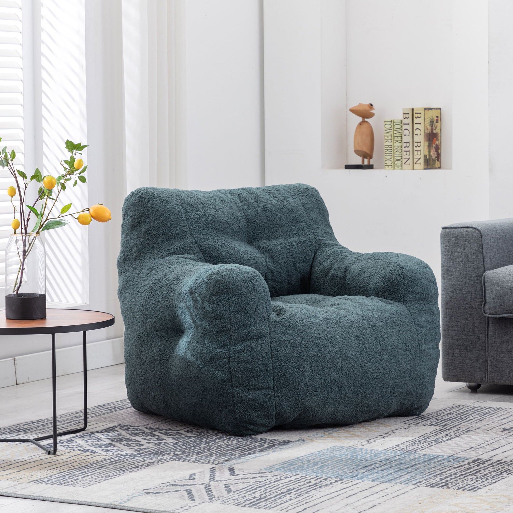 A Soft Teddy Fabric Tufted Foam Bean Bag Chair sits on a patterned rug in a bright living room with a gray sofa, a round table topped with yellow lemons, and a bookshelf holding books and a wooden sculpture.