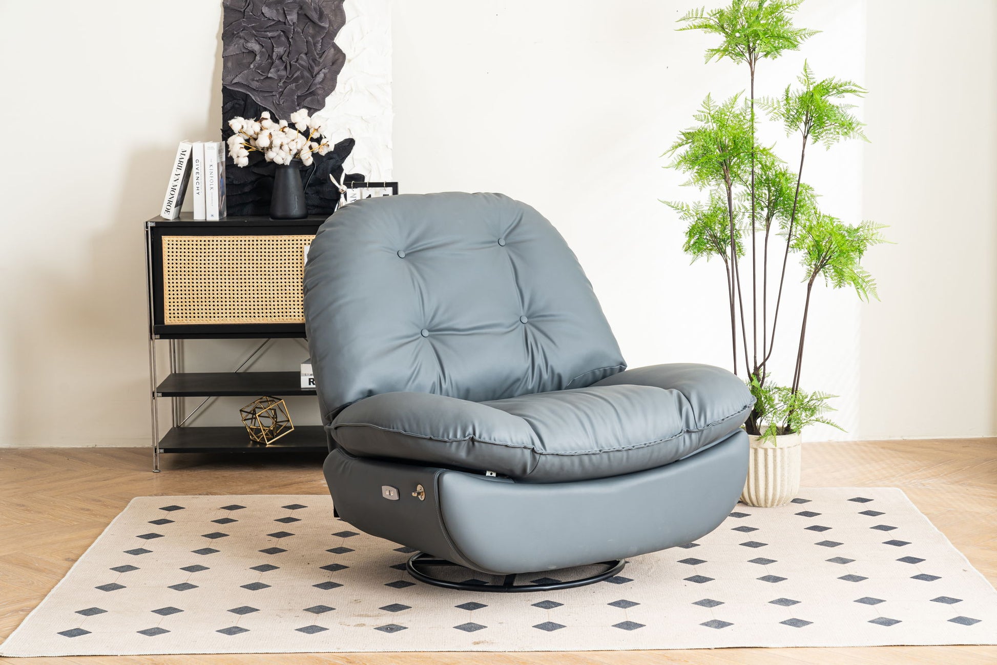A gray Power Recliner Swivel Glider with USB charger and Bluetooth music player sits on a light rug with black diamond patterns, near a black shelf of books and decor, and a tall green potted plant against a white wall.