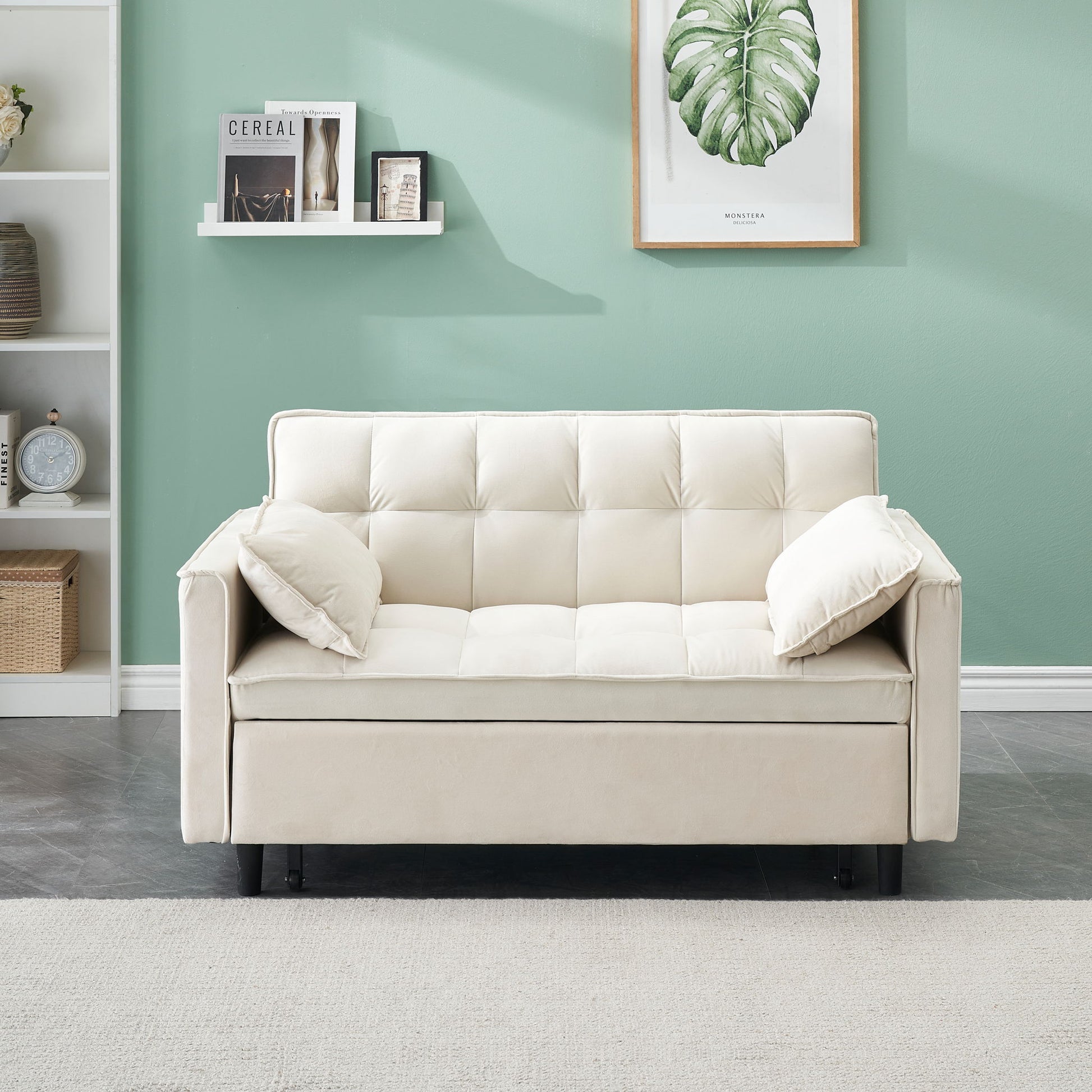 A Modern Velvet Sofa pull-out bed with pillows and pockets sits against a mint green wall below a framed leaf print. Nearby, a white shelf holds books and decor, and a beige rug lies on the floor.