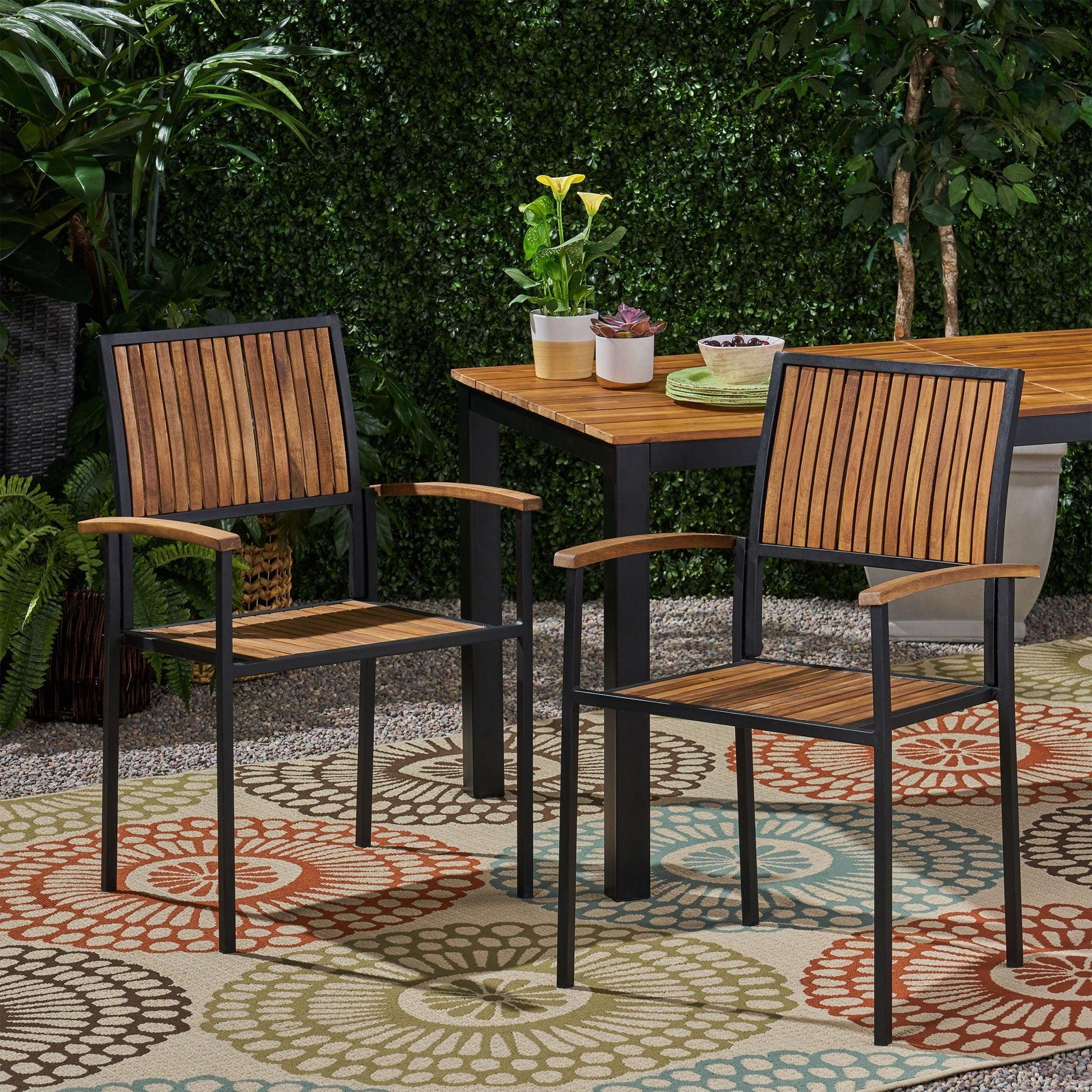 Two Vermilion Outdoor Dining Chairs with vertical slats and wooden arms sit on a vibrant outdoor rug beside a matching table, surrounded by greenery and potted plants.