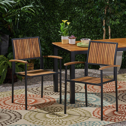 Two Vermilion Outdoor Dining Chairs with vertical slats and wooden arms sit on a vibrant outdoor rug beside a matching table, surrounded by greenery and potted plants.