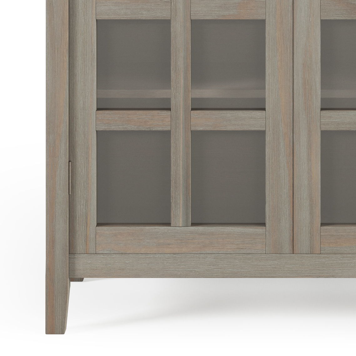 Close-up of the lower corner of the Acadian - Medium Storage Cabinet, Handcrafted, featuring a light weathered wood finish, glass panel door, and short legs.