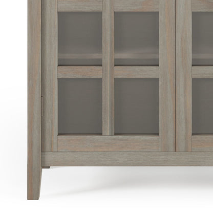 Close-up of the lower corner of the Acadian - Medium Storage Cabinet, Handcrafted, featuring a light weathered wood finish, glass panel door, and short legs.