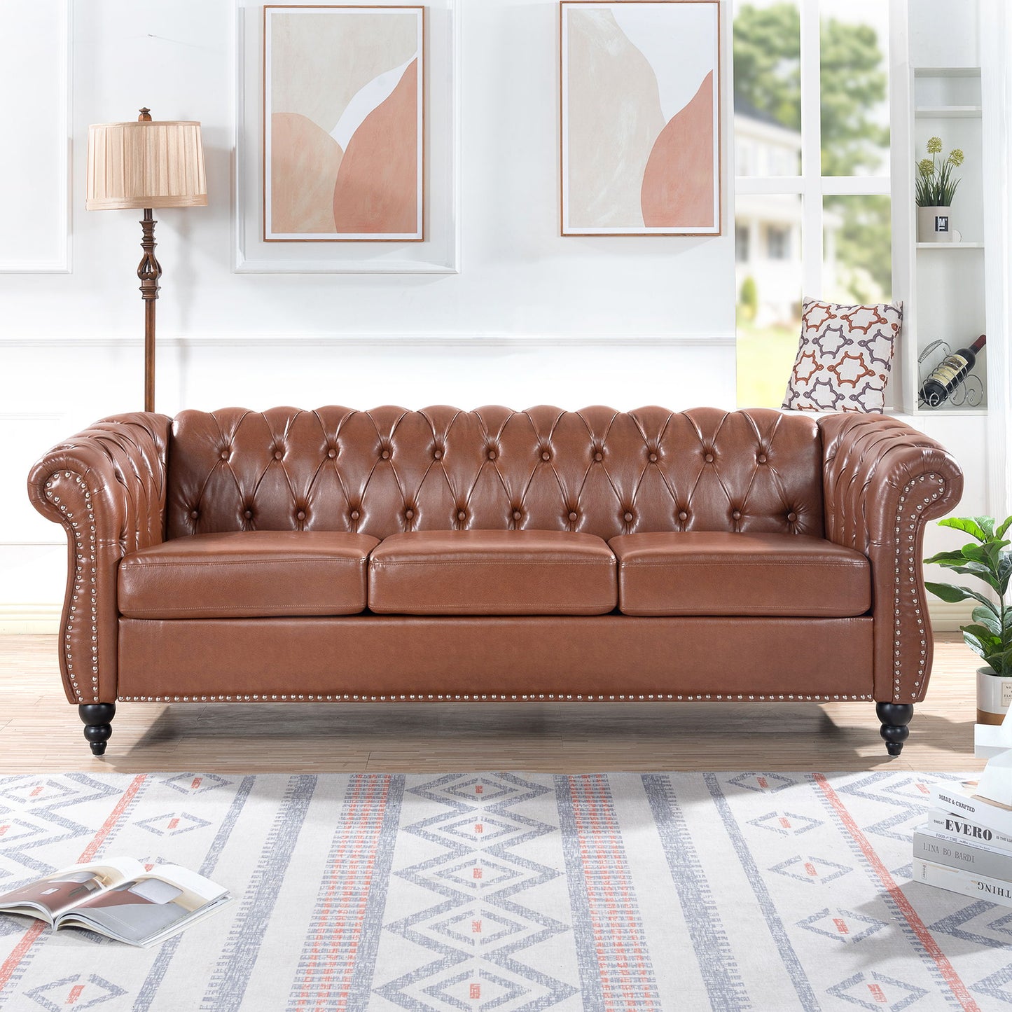 The Rolled Arm Chesterfield 3 Seater Sofa, upholstered in brown tufted leather, stands in a bright living room with framed abstract art, a floor lamp, bookshelves, and a patterned rug.
