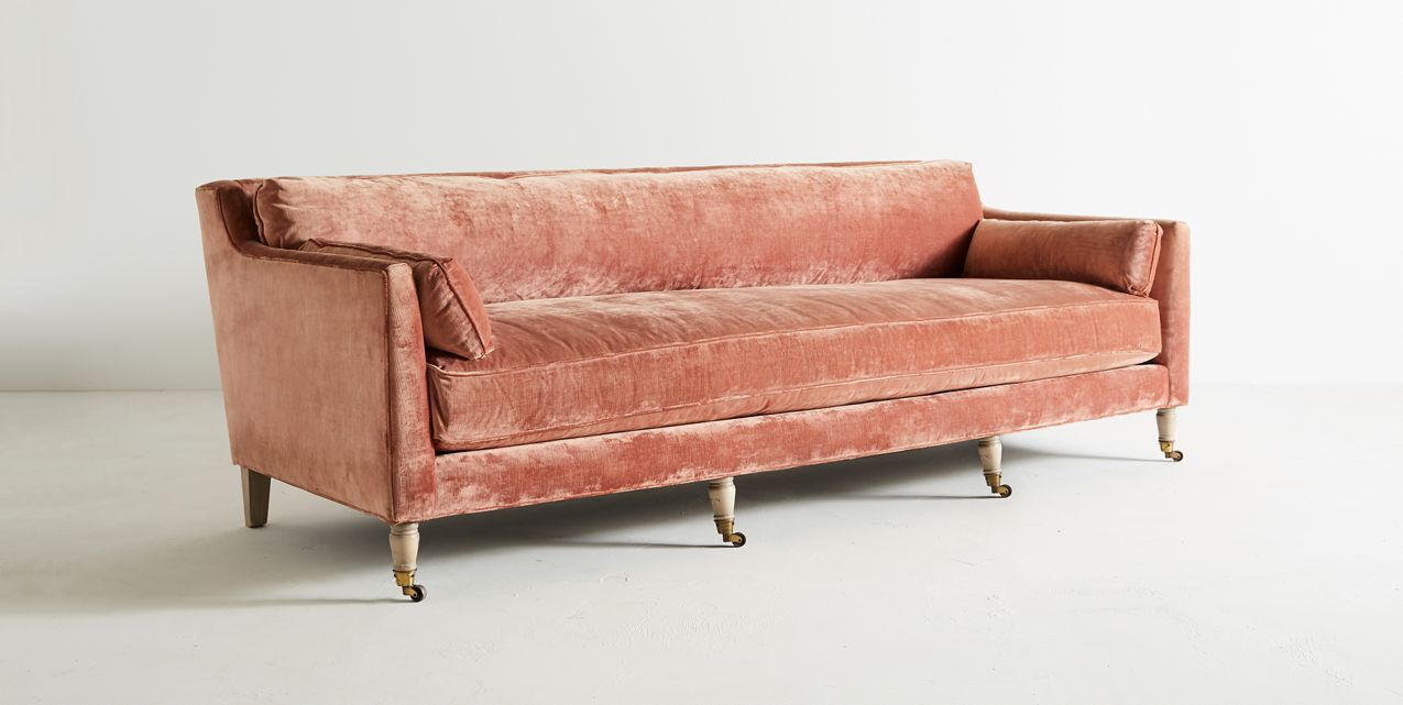 The Leonelle Sofa, a vintage-style piece in pink velvet with wooden legs on small wheels, sits against a plain white background. It features a plush seat cushion and padded armrests, embodying a classic and elegant design.