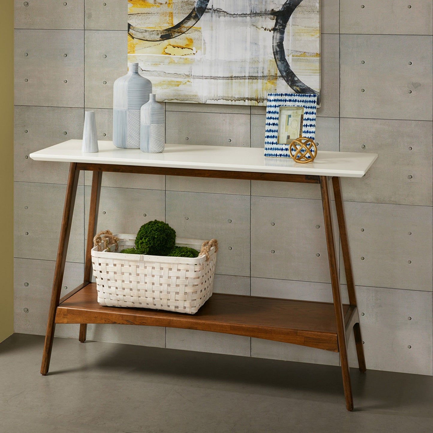 The Modern Table With Extra Storage features a white top and wooden legs displaying vases, decor, and a picture frame. A woven white basket with green spheres sits on the lower shelf beneath a geometric painting on the gray wall.