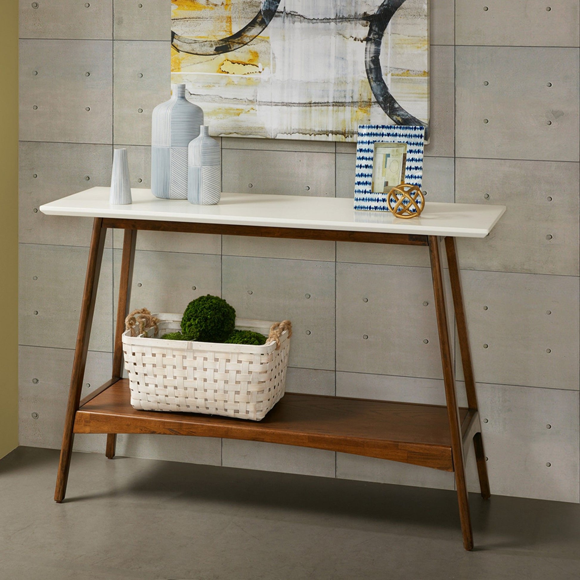 The Modern Table With Extra Storage features a white top and wooden legs displaying vases, decor, and a picture frame. A woven white basket with green spheres sits on the lower shelf beneath a geometric painting on the gray wall.