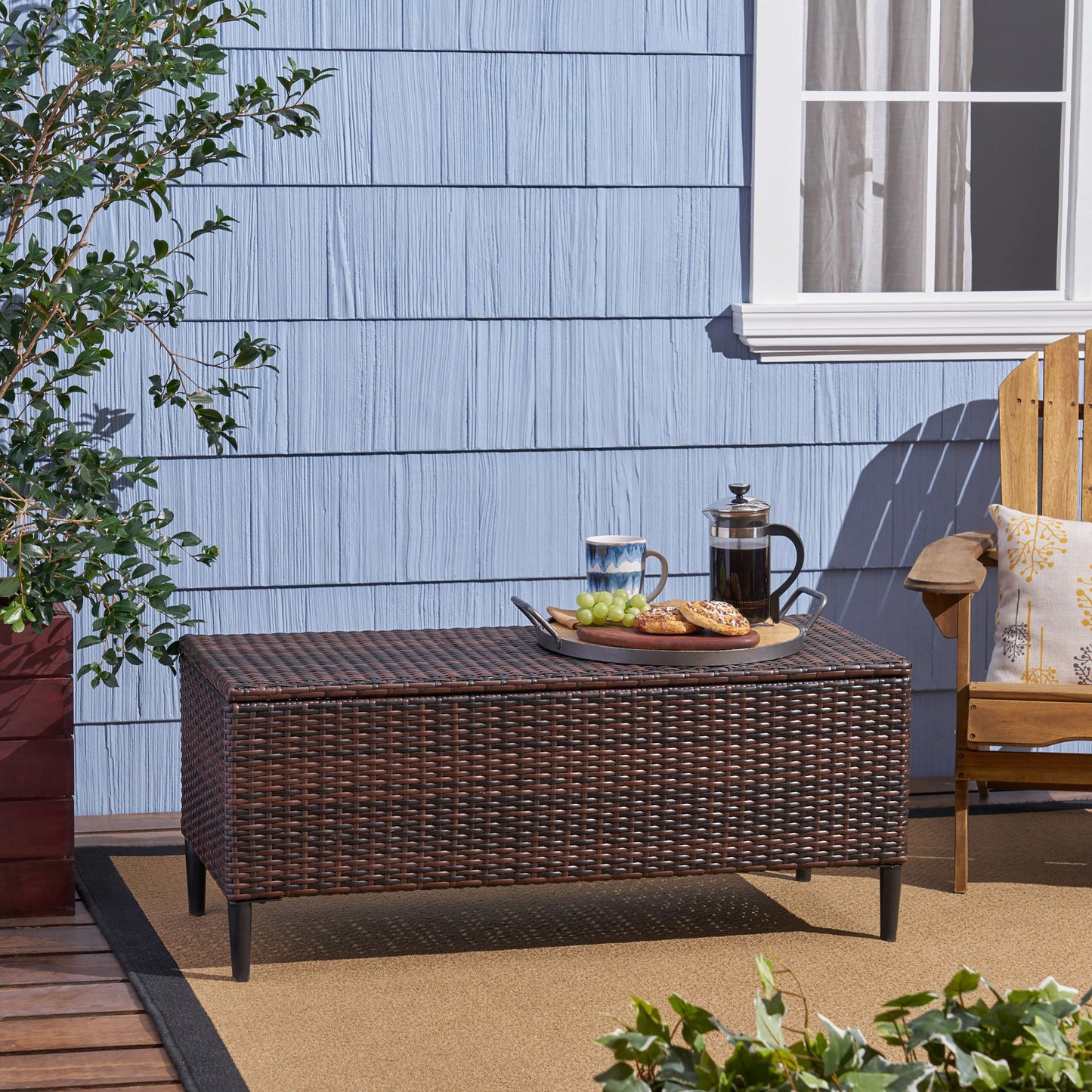 The Rupert - Storage Unit Resin Wicker Outdoor sits on a patio with a blue paneled wall and window behind it. On top are a tray, coffee press, glass cup, grapes, and pastries; nearby are a wooden chair and potted plant.