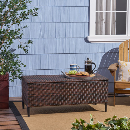 The Rupert - Storage Unit Resin Wicker Outdoor sits on a patio with a blue paneled wall and window behind it. On top are a tray, coffee press, glass cup, grapes, and pastries; nearby are a wooden chair and potted plant.