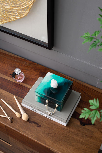 The Bethany - Aqua Jewelry Box, a decorative green box, sits on stacked books on a wooden table near a small perfume bottle, wooden utensils, and a leafy plant in the corner.
