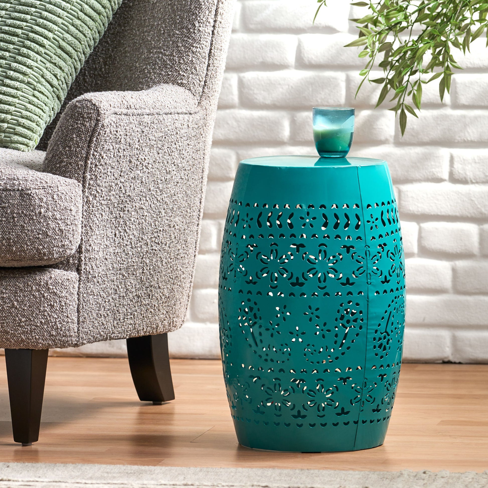 A 12" Outdoor Lace Cut Side Table in teal, featuring intricate patterns and a small green candle on top, stands next to a gray upholstered chair on a wooden floor with a white brick wall and green plant behind.