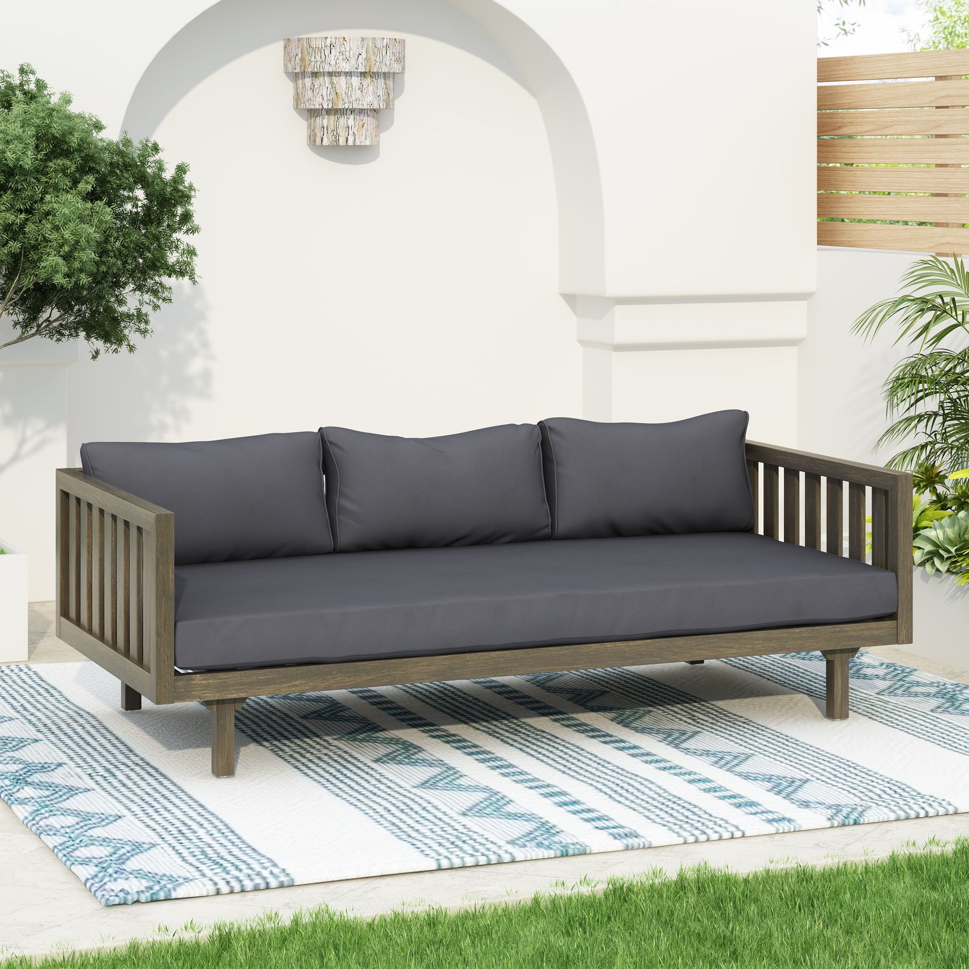 The Claremont 3 Seater Daybed, made from teak acacia wood with water-resistant dark gray cushions, is placed on a blue and white patterned rug in a garden patio with greenery and white arched walls.
