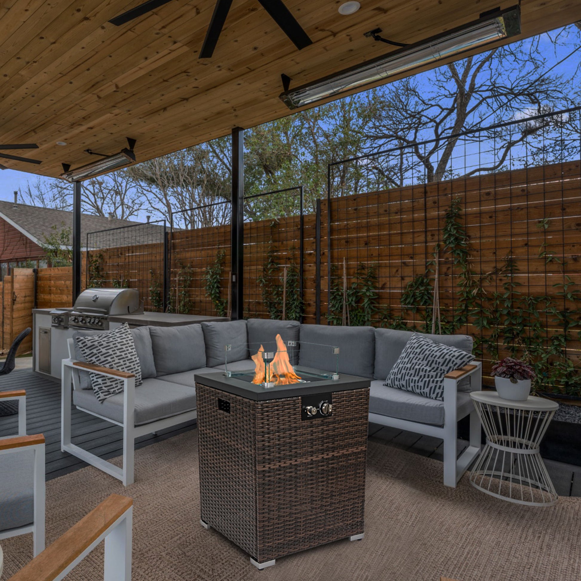 A cozy outdoor patio features a gray sectional sofa, a Propane Gas Square Fire Pit with tile tabletop and wicker base, a grill, wooden privacy fence, climbing plants, and a wooden pergola ceiling with fans.