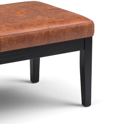 A close-up of the Lacey - Upholstered Tufted Ottoman Bench shows its brown leather seat and black wooden legs, with only the corner of the seat and two legs visible against a white background.