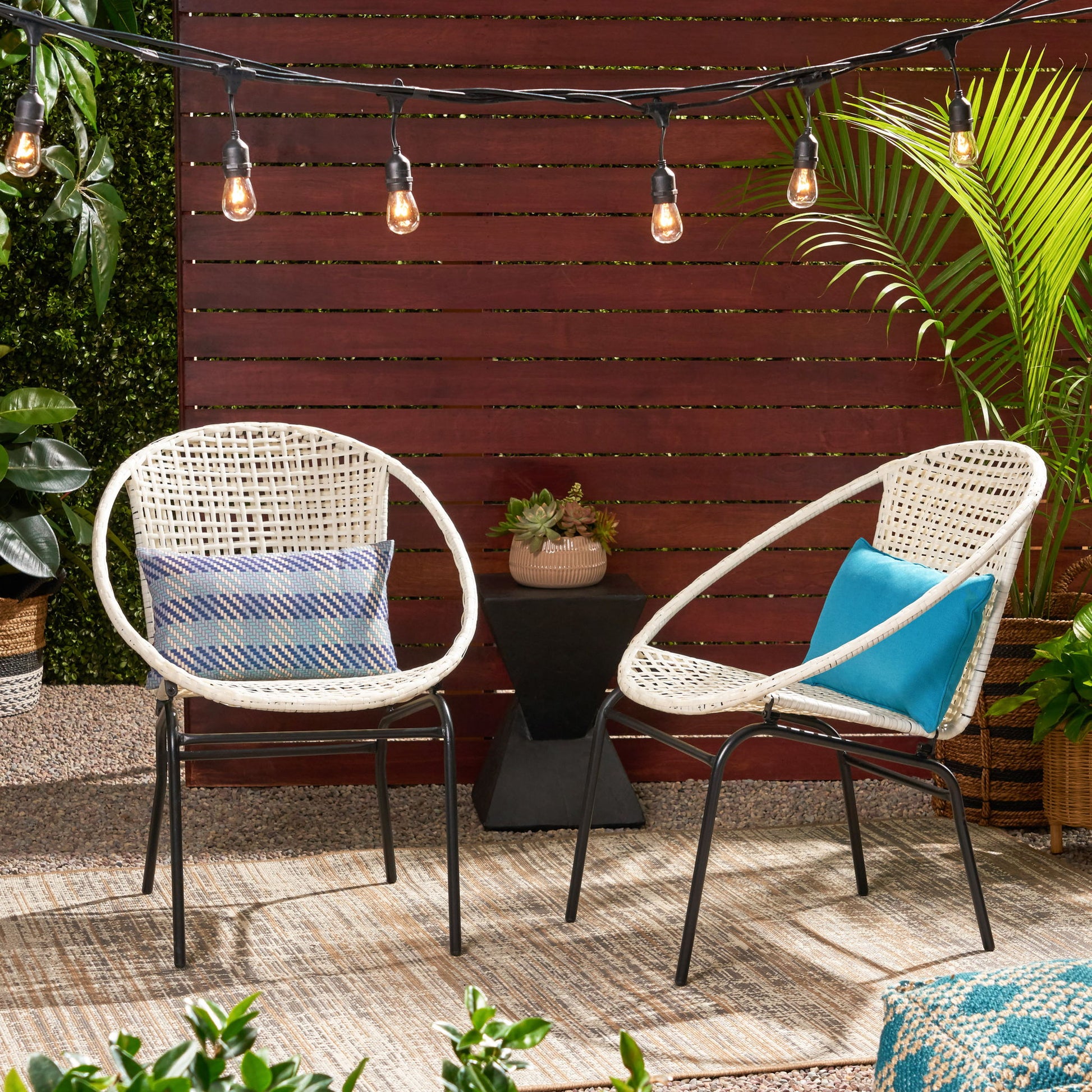 Two Java Chairs With Comfortable Design and cushions sit on a rug outdoors. String lights are above, with potted plants and a wooden privacy fence in the background.