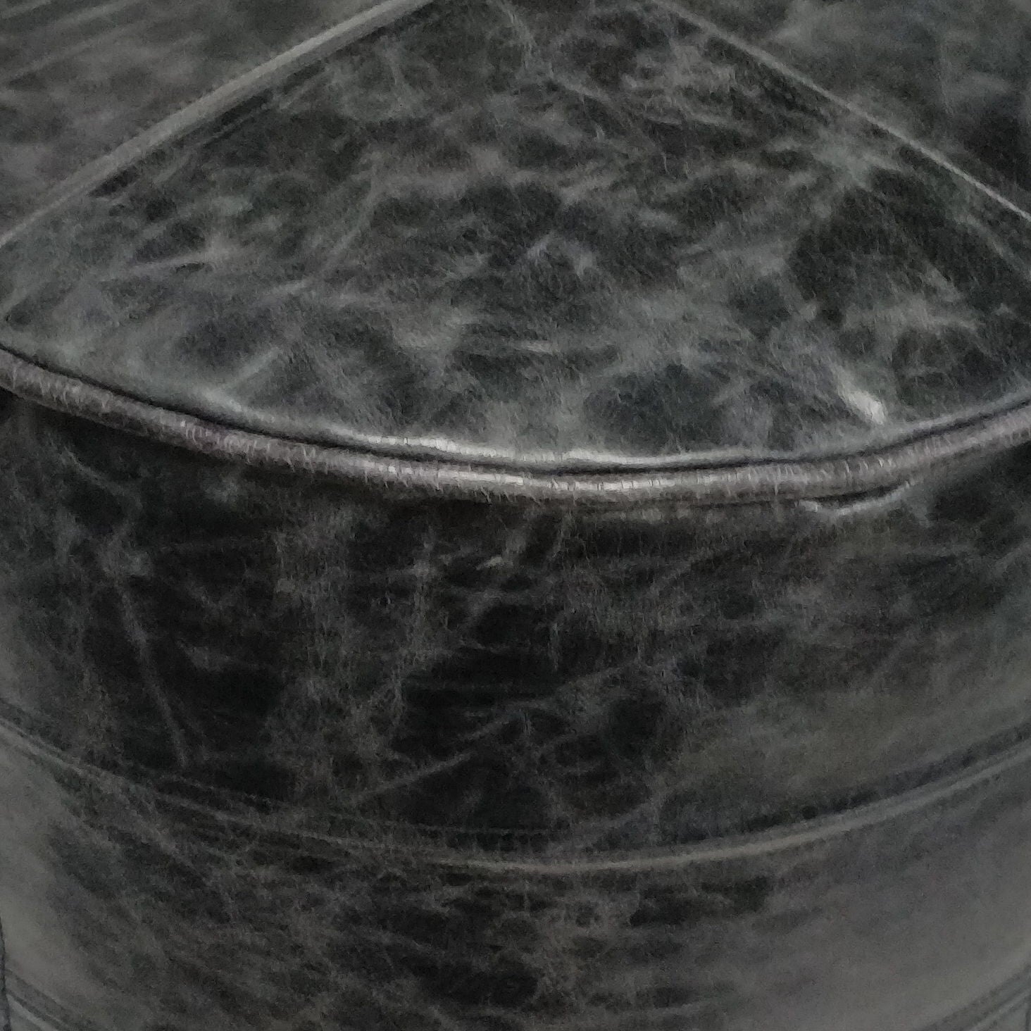 Close-up of the Connor Handcrafted Round Pouf's black leather surface, highlighting creases, stitching, a rounded edge, and its textured, worn appearance.