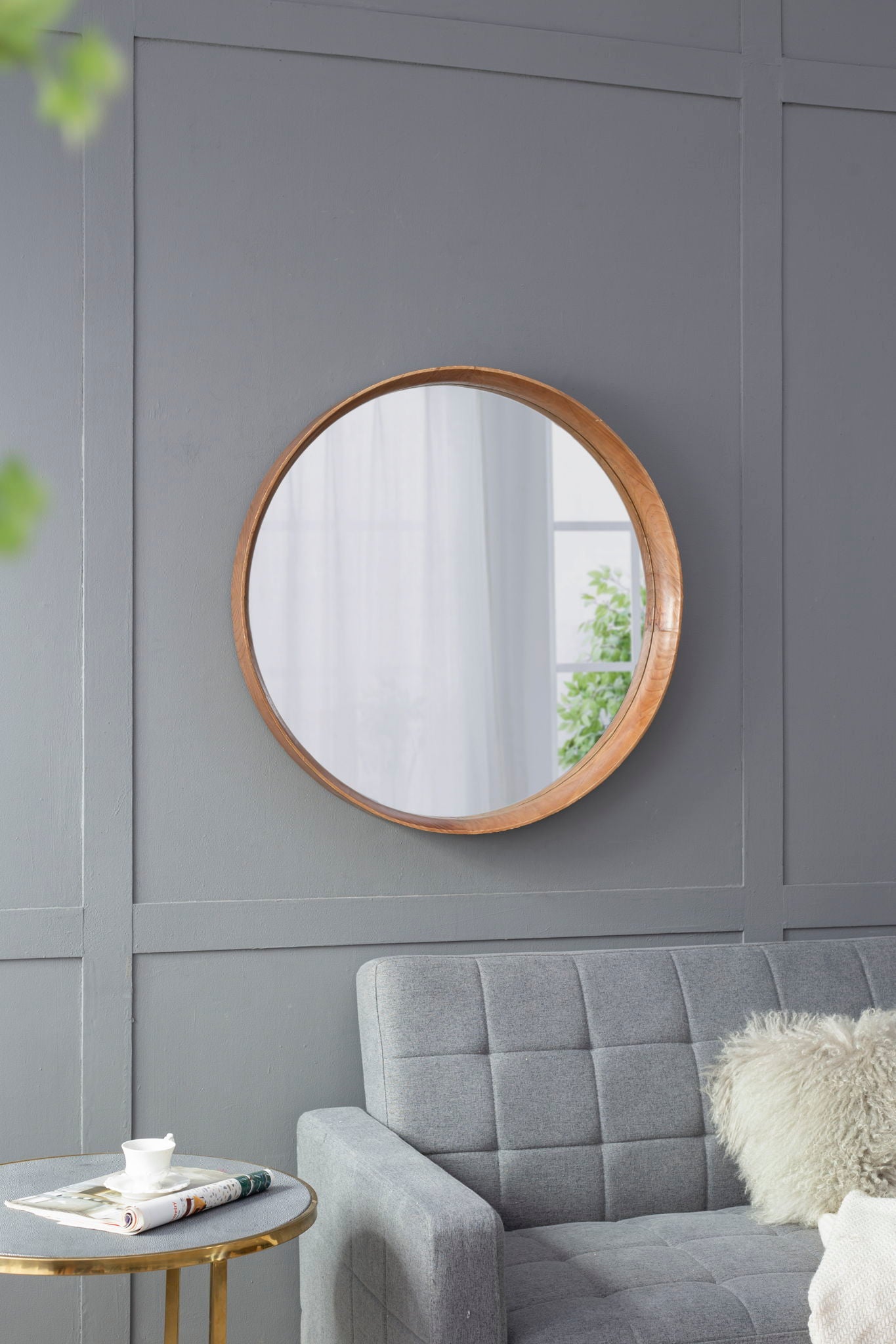 A Round Wood Mirror, Wall Mounted Mirror Home Decor For Bathroom Living Room, hangs on a gray paneled wall above a light gray tufted sofa with a gold side table holding a teacup, saucer, and magazines; greenery is reflected in the mirror.