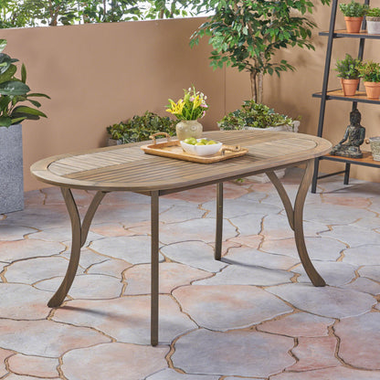 An Outdoor Table With Smooth Lines, Slat Top sits on a tiled patio, topped with a tray, bowl, and vase of flowers. Potted plants and a small statue decorate the nearby wall and black shelf.