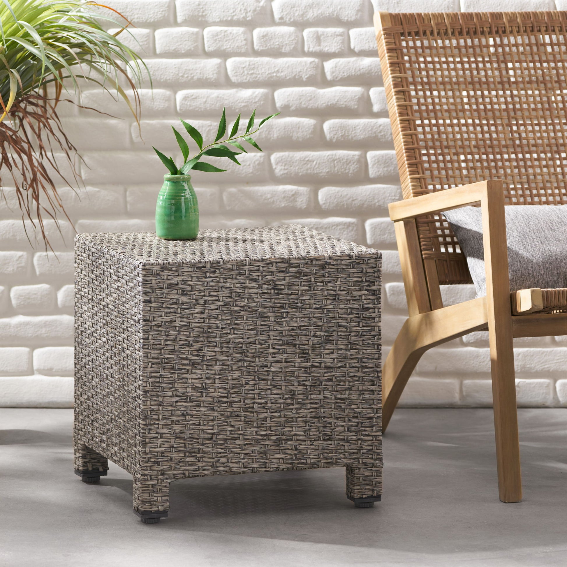 The Puerta Minimalist Style Accent Table with a small green vase sits beside a wooden and rattan armchair with a gray cushion, set indoors against a white brick wall with a nearby potted plant.