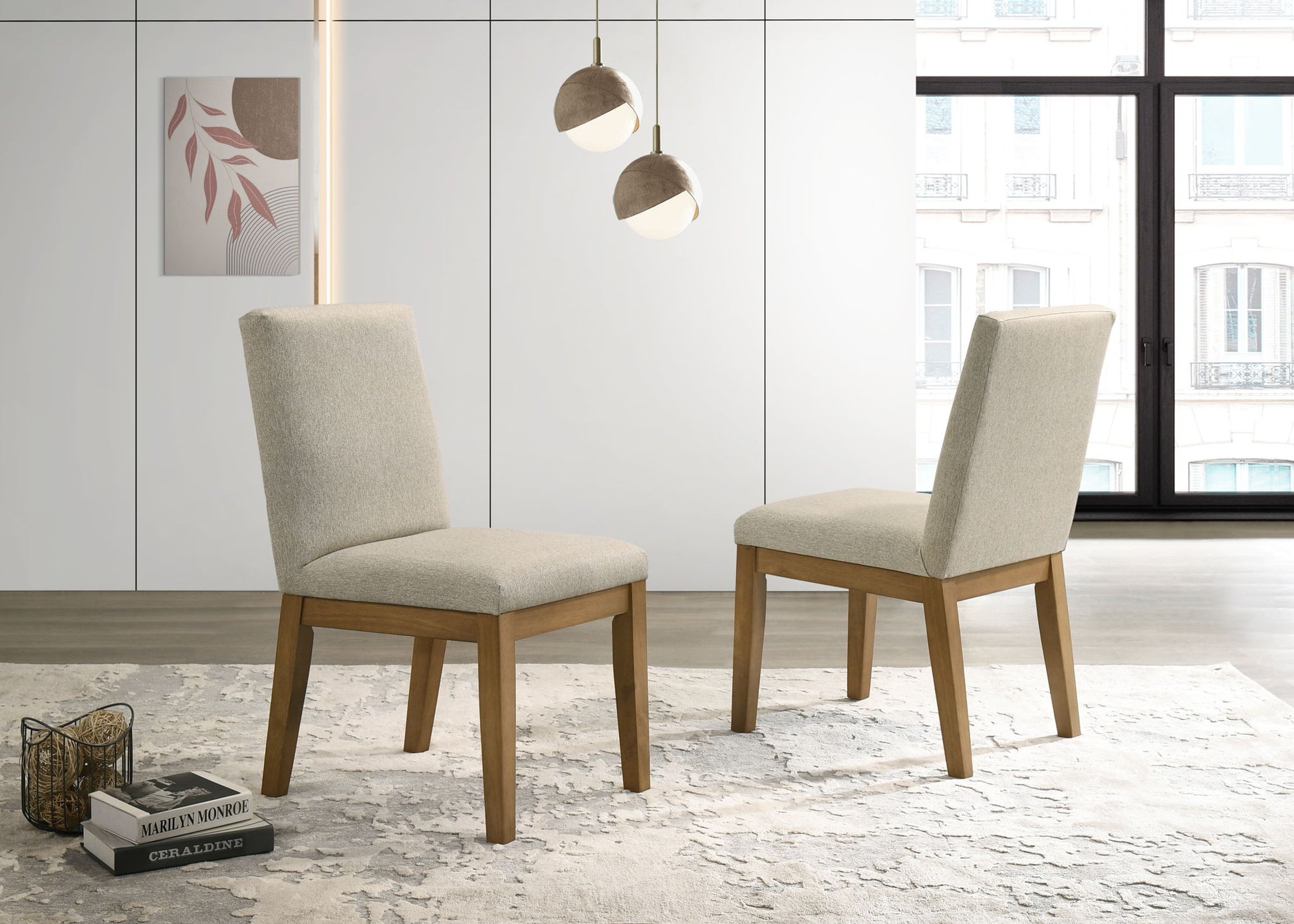 Two Jasper - Contemporary Fabric 19" Dining Chairs (Set of 2), upholstered in beige with wooden legs, are set on a light patterned rug in a modern room featuring white cabinets, large windows, pendant lights, and wall art.