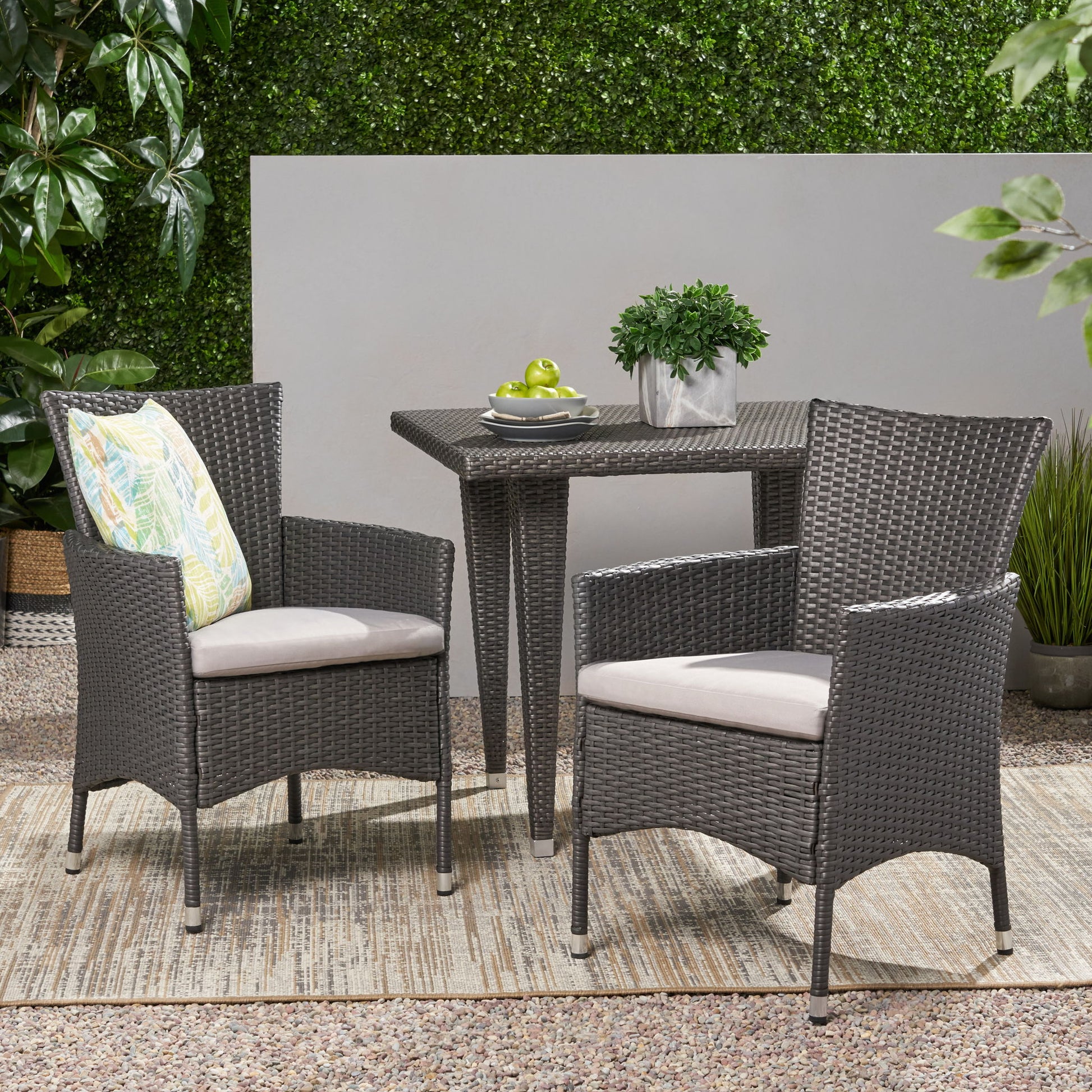 Two Dimensional Outdoor Dining Chairs with cushion seats and iron frames (set of 2) sit beside a square wicker table with a plant and apples, all set on an outdoor rug with a green hedge in the background.