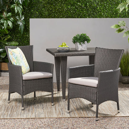 Two Dimensional Outdoor Dining Chairs with cushion seats and iron frames (set of 2) sit beside a square wicker table with a plant and apples, all set on an outdoor rug with a green hedge in the background.