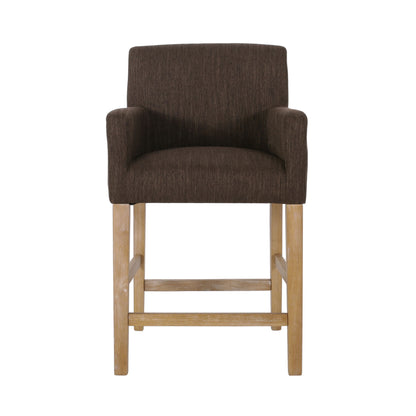 The Charmed Upholstered Counter Stool features round arms, a cushioned back, brown upholstery, light wooden legs with an H-frame base and footrest, shown from the front against a white background.