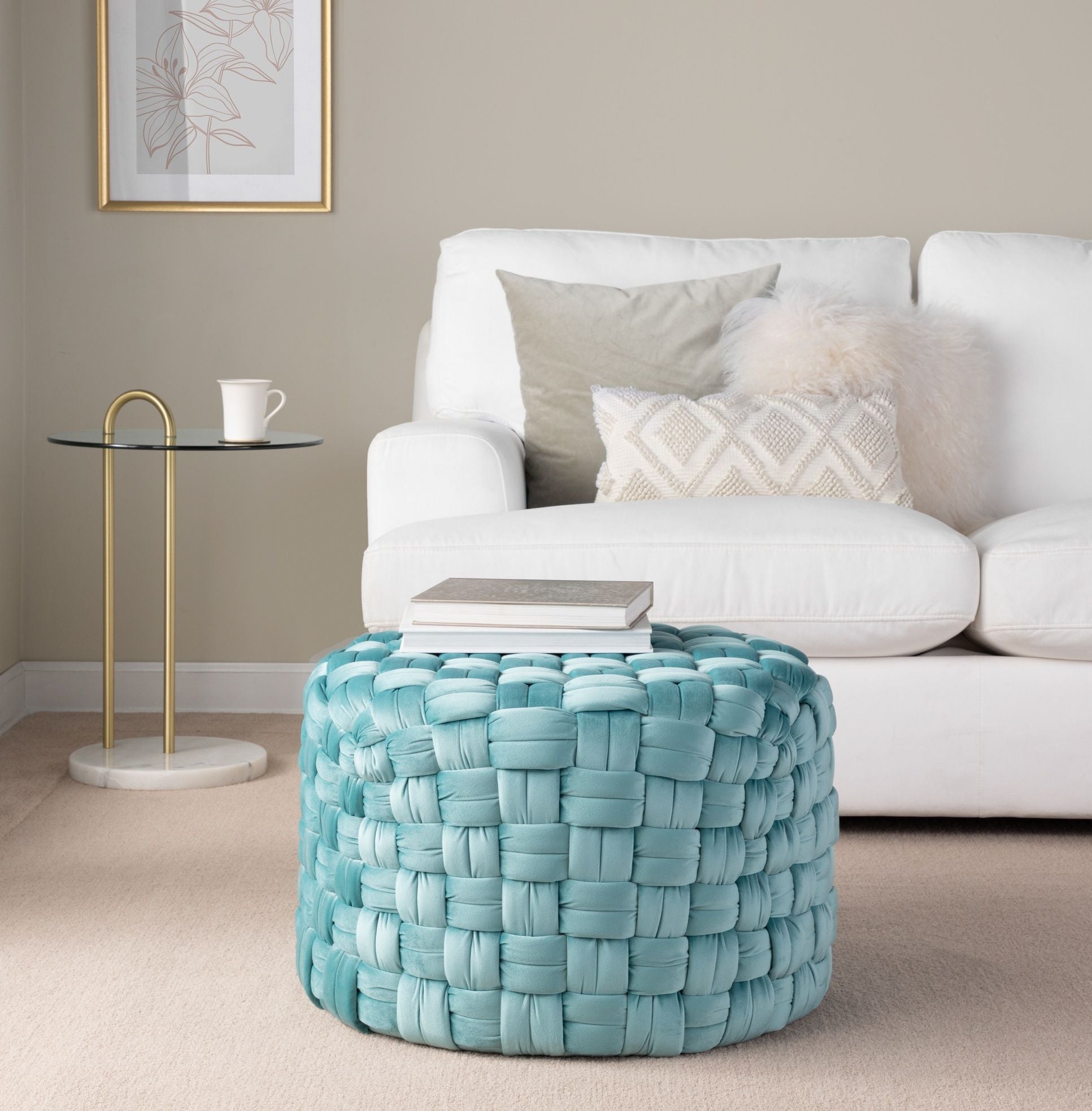 A cozy living room with a white sofa, light pillows, a fluffy cushion, a round glass side table with a cup, and the Braided - Versatile Round Ottoman in blue topped with stacked books on beige carpet.