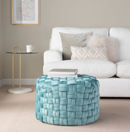 A cozy living room with a white sofa, light pillows, a fluffy cushion, a round glass side table with a cup, and the Braided - Versatile Round Ottoman in blue topped with stacked books on beige carpet.